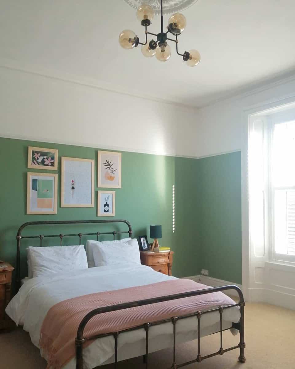 Bedroom with a metal bed, green walls, framed art, and a chandelier illuminated by natural light from a large window