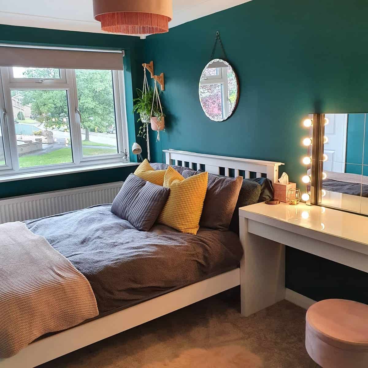 Bedroom with teal walls, a bed with gray and yellow pillows, a round mirror, and a makeup vanity by the window