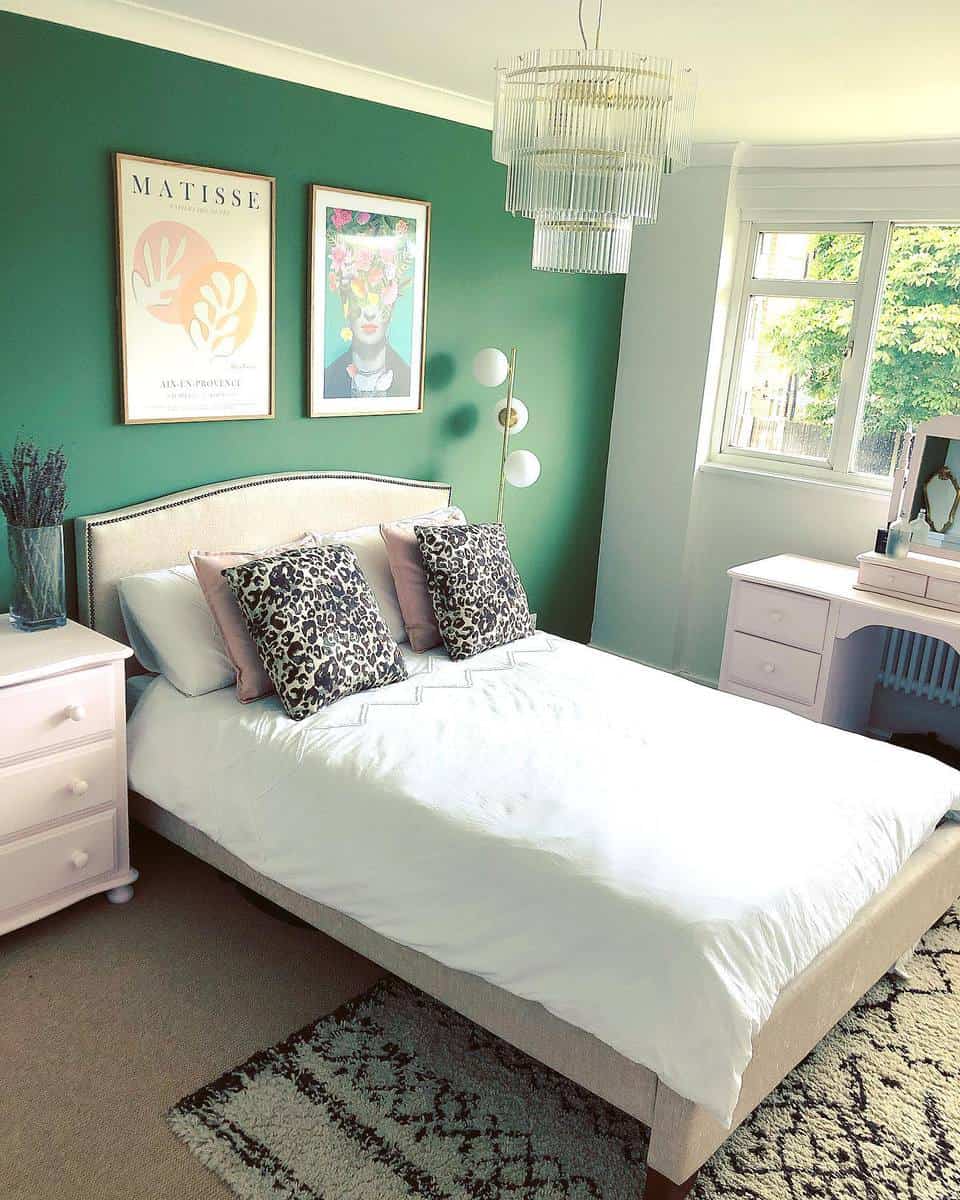 Stylish bedroom with a green accent wall, artwork, a white bed, and a chandelier next to a window with a garden view