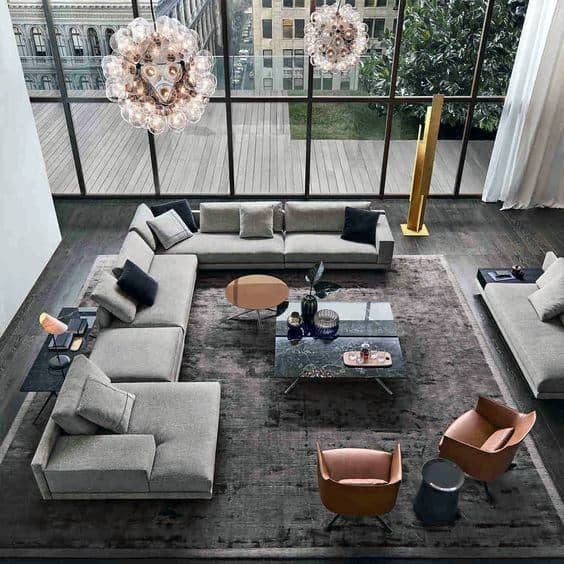 Modern living room with gray sectional sofa, glass coffee table, two chandeliers, and floor-to-ceiling windows