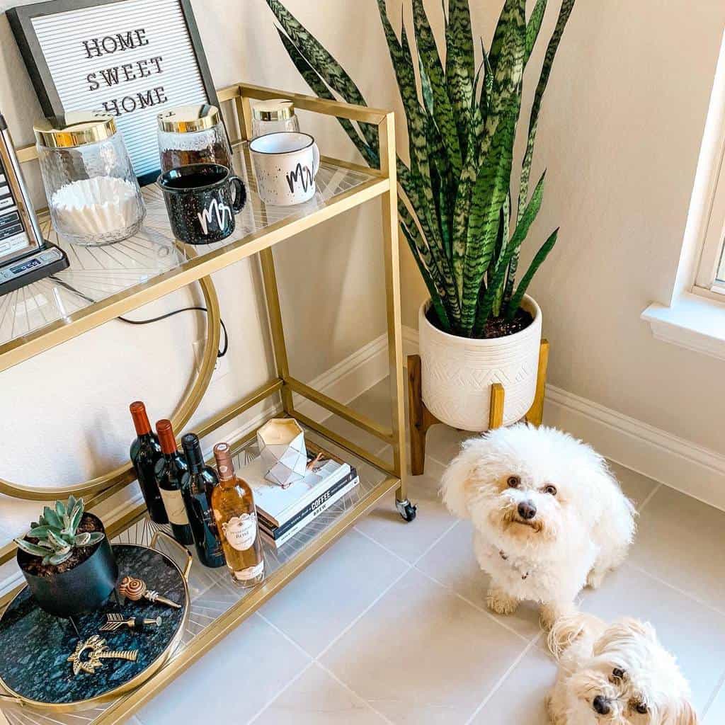 Gold glass bar cart with mugs, wine bottles, books, and two small white dogs nearby.