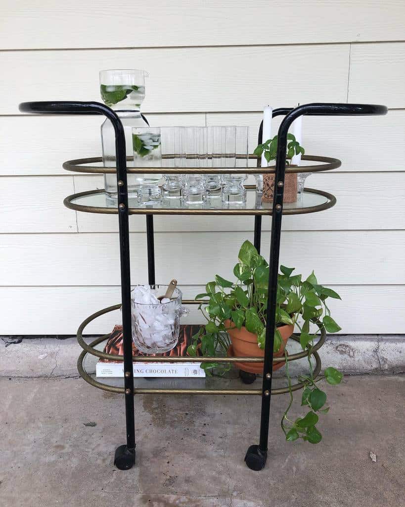 Gold and black cocktail bar cart with glassware, a water pitcher, ice bucket, and potted plant.