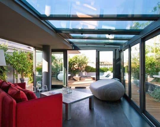 A modern sunroom with a glass roof, sleek black-framed glass walls, a bold red sofa, and minimalist furnishings.