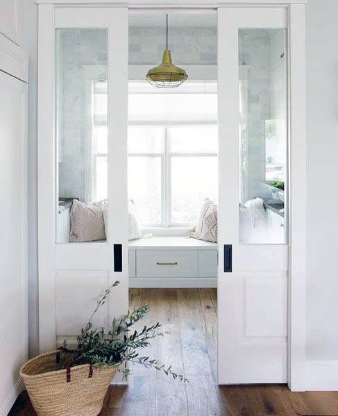 Elegant white pocket doors with glass panels open to a cozy window nook featuring built-in seating, pillows, and natural light