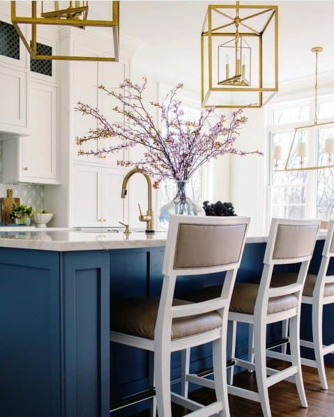 Charming kitchen with gold pendant fixtures above a blue island, complemented by white bar stools and a beautiful floral arrangement