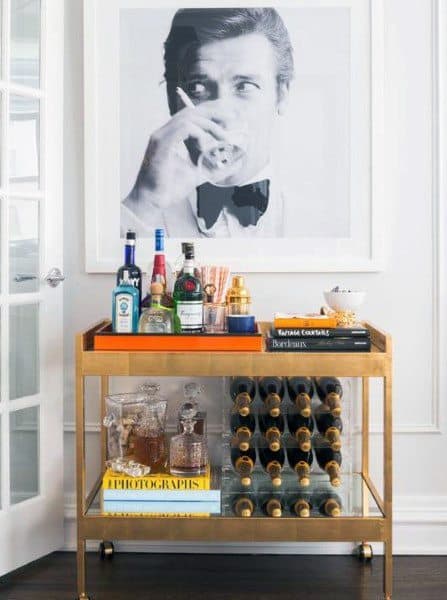 Chic gold bar cart with liquor bottles, glassware, wine storage, and a stylish wall portrait.