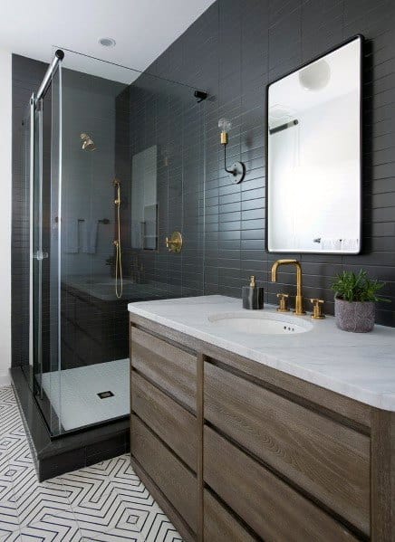 Modern bathroom with black subway tile walls, gold fixtures, wood vanity, and patterned floor tiles.