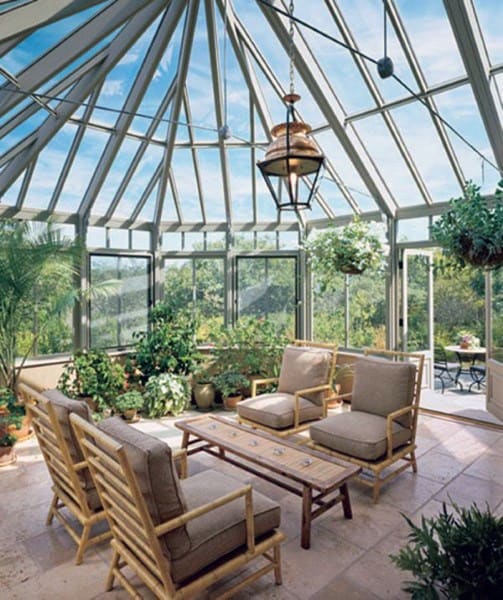 The sunroom features a vaulted glass ceiling, bamboo furniture with beige cushions, and abundant greenery for a natural, airy look.