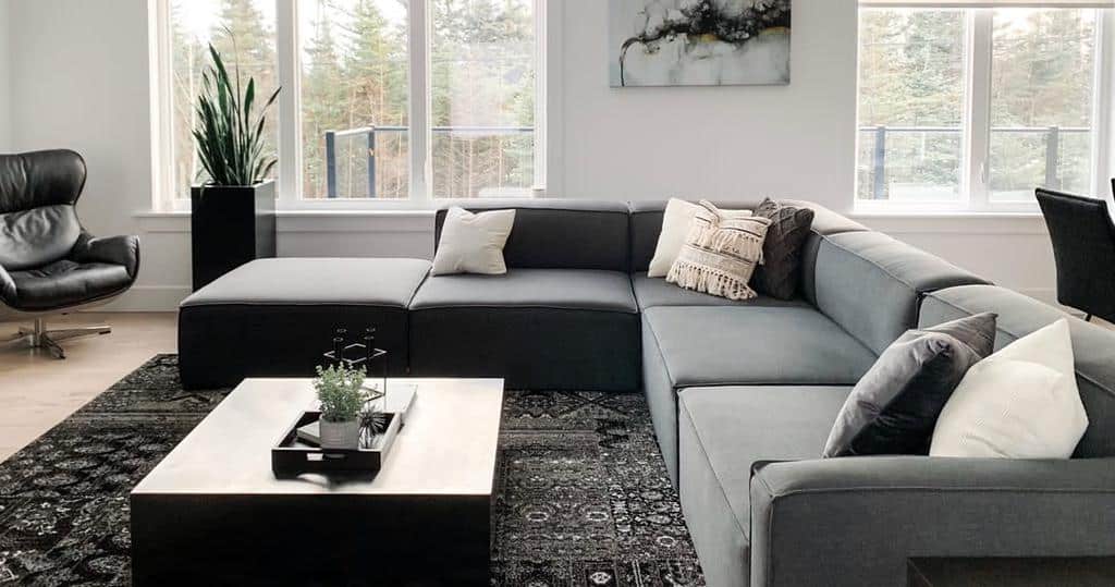 Modern living room with a gray sectional sofa, black leather chair, patterned rug, and large windows with forest views