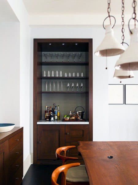 Compact mini bar with dark wood shelves, glassware, and a white countertop in a dining area.