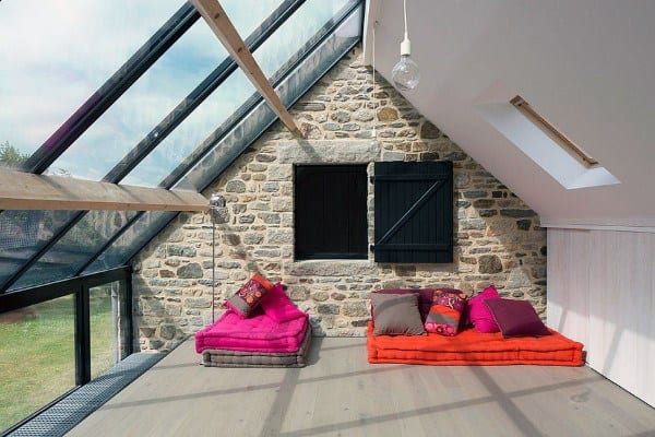 Sunroom with a rustic stone wall, slanted glass ceiling, and vibrant floor cushions for a cozy, relaxed atmosphere.