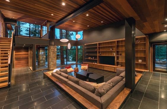 Modern living room with wooden ceiling, large windows, black tile floor, and a sectional sofa facing a TV on shelves