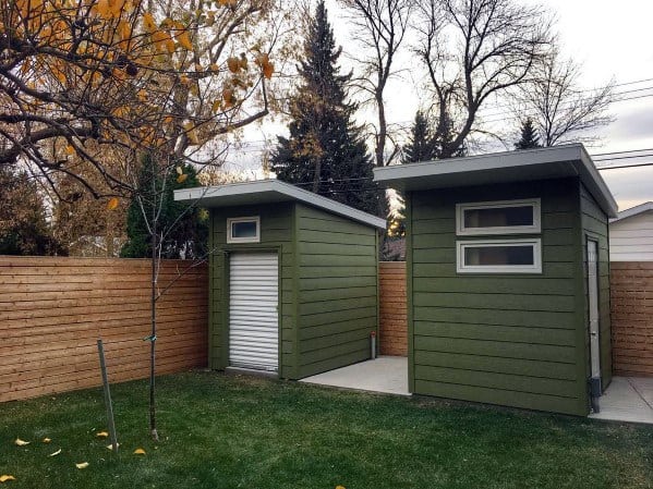 Two green pre-fabricated vinyl sheds with modern flat roofs in a fenced backyard.