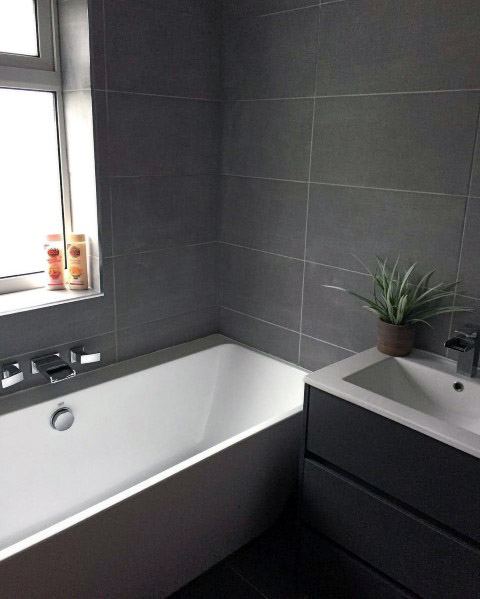 Modern bathroom with a grey touch, featuring a white bathtub, sink, plant, and bottles by the window