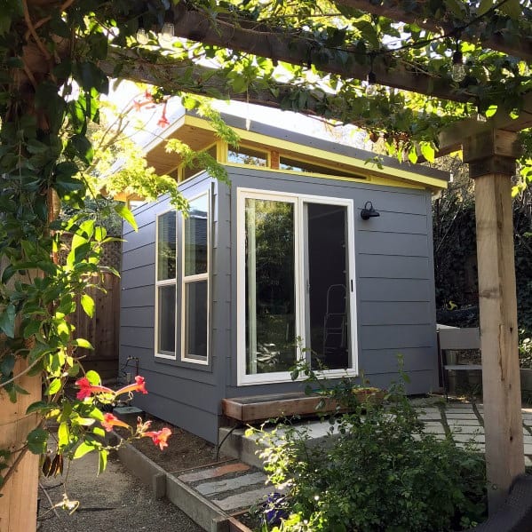 Gray pre-fabricated vinyl shed with large windows and sliding glass door, surrounded by greenery.