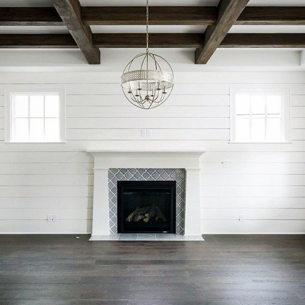 A farmhouse-style fireplace featuring gray patterned tiles framed by a white shiplap wall and rustic wooden beams overhead.