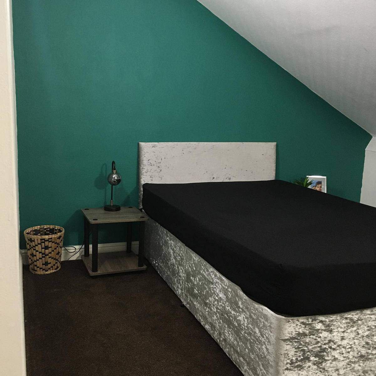 Minimalist bedroom with a teal accent wall, a single bed, a side table with a lamp, and a woven basket