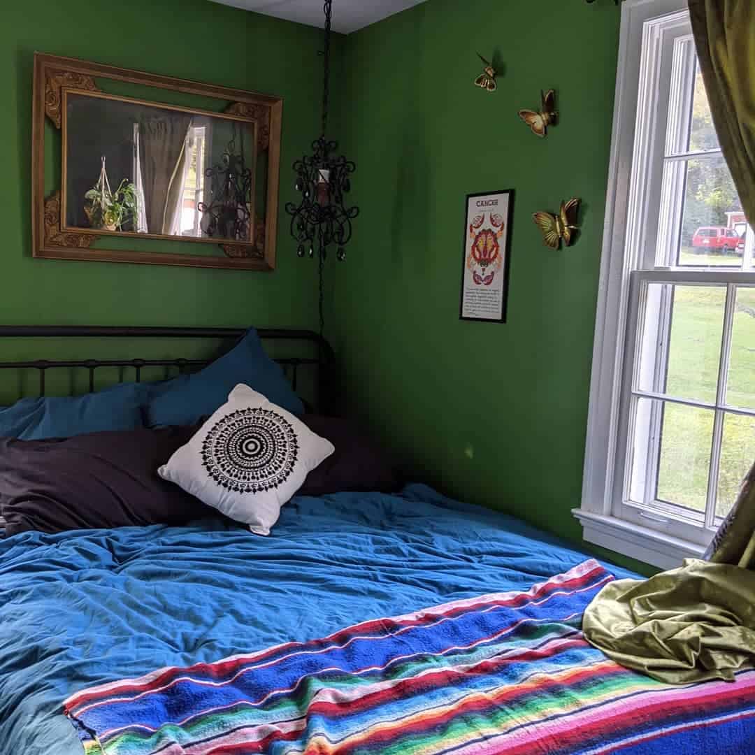 Cozy bedroom with green walls, a bed with colorful blankets, decorative pillows, and a window with greenery outside