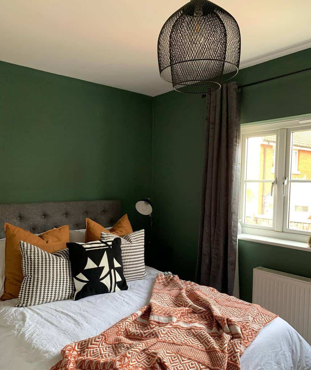 Cozy bedroom with green walls, modern pendant light, patterned pillows, and a window with gray curtains