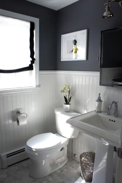 Modern bathroom with dark walls, white wainscoting, toilet, and sink, window with blinds and framed art above toilet