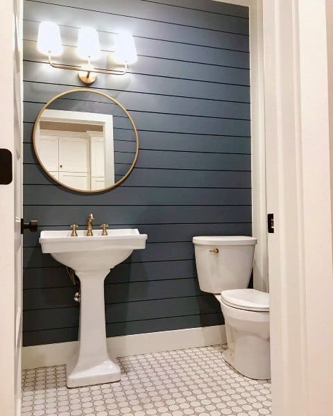 Half bath with blue shiplap walls, round mirror, white sink, toilet, and hexagonal tile flooring under warm lighting