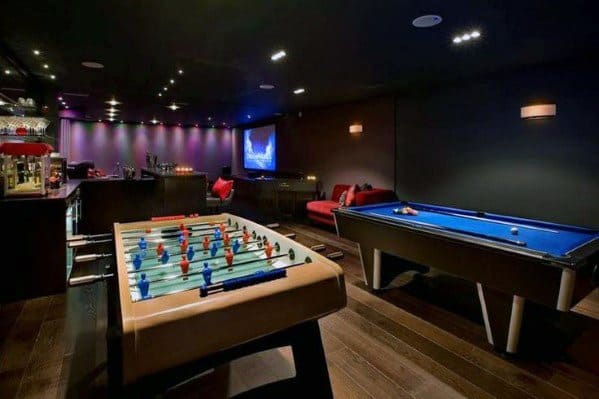 A game room featuring a foosball table, pool table, and bar area with dim lighting and a large screen