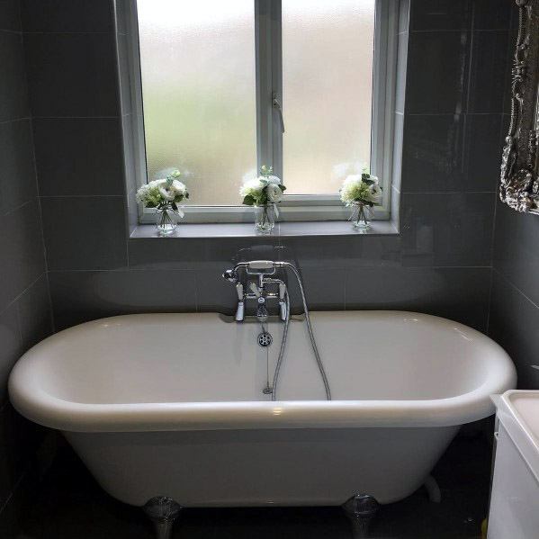 White bathtub in a gray-tiled bathroom with a window and floral arrangements