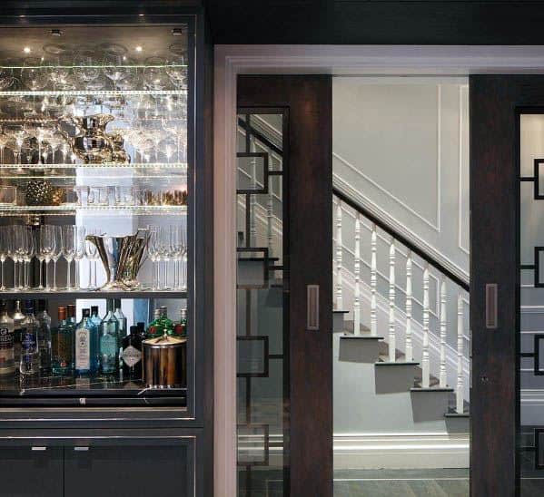 Elegant dark wood pocket doors with glass and geometric detailing open to a stylish staircase, next to a mirrored bar cabinet