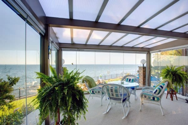 A bright sunroom with glass walls, wicker chairs, striped cushions, and lush green plants overlooking a scenic view.