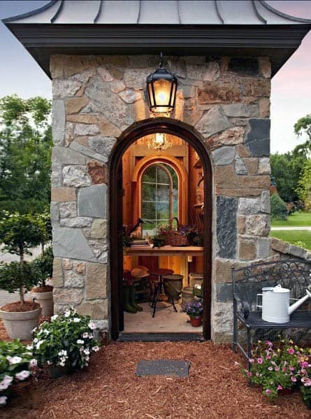 Charming stone shed with arched doorway, lantern light, and cozy gardening workspace inside.