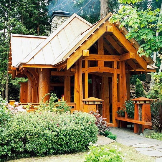 Large wooden garden shed with intricate beams and a covered porch, surrounded by greenery.