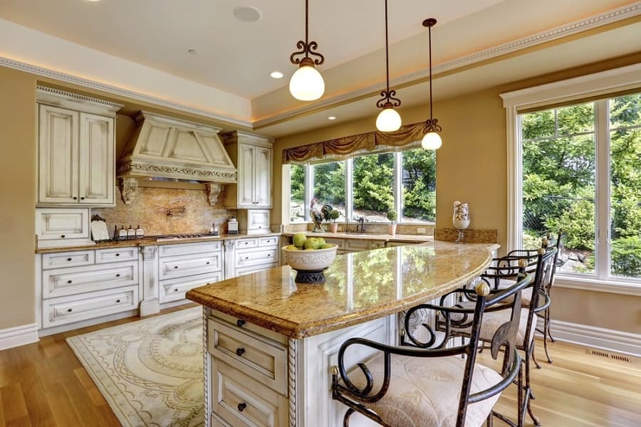 Elegant kitchen with a granite island, high chairs, white cabinets, large windows, and sleek hanging lights