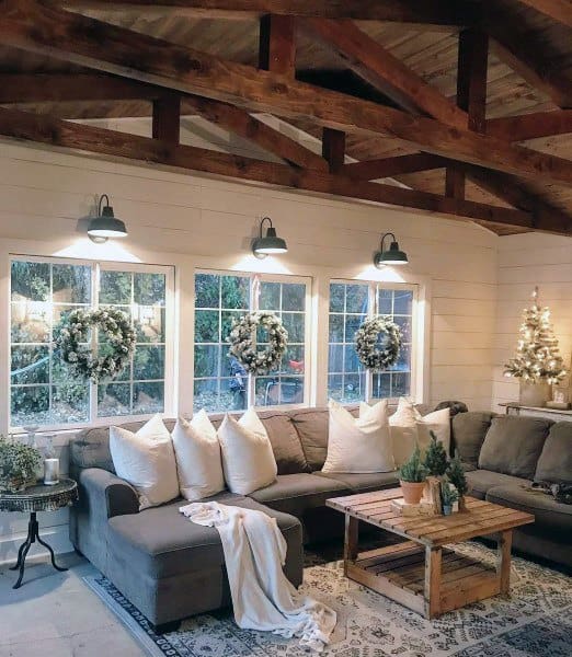 A cozy rustic living room with exposed wooden beams, large windows adorned with wreaths, and a comfy sectional sofa, creating a warm and inviting atmosphere