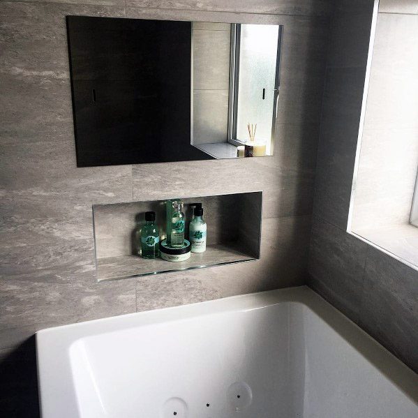 Modern bathroom with grey tiled walls, built-in shelf displaying toiletries, a window with natural light, and a white bathtub