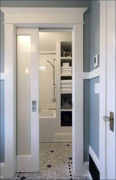 White pocket door with glass panel, framed in wood, leads to a bathroom with built-in shelves and a tiled shower