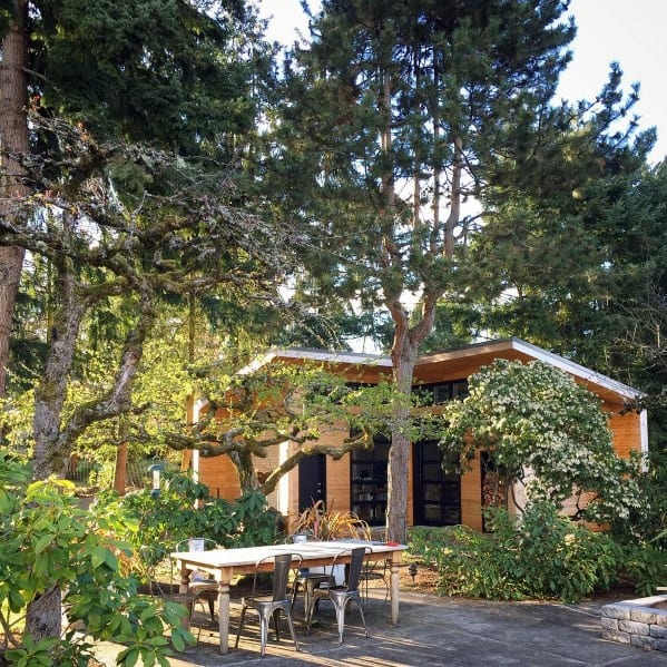 Modern wooden shed surrounded by lush greenery with an outdoor dining table under trees.