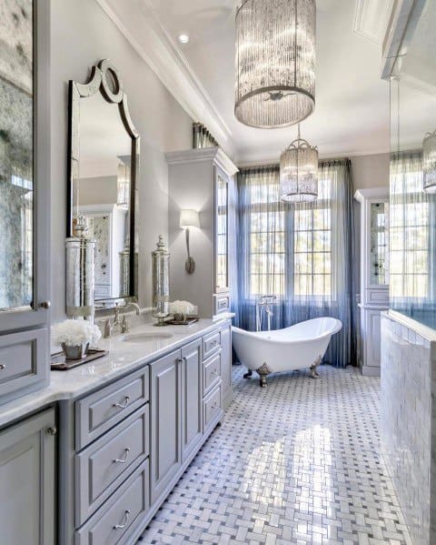 Sophisticated bathroom with freestanding clawfoot tub, double chandeliers, and gray cabinetry.