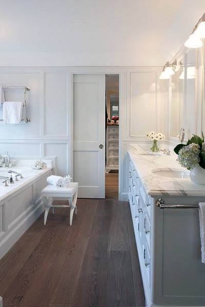 Bright white bathroom with a classic pocket door, marble countertops, double sinks, a built-in tub, and elegant panel detailing