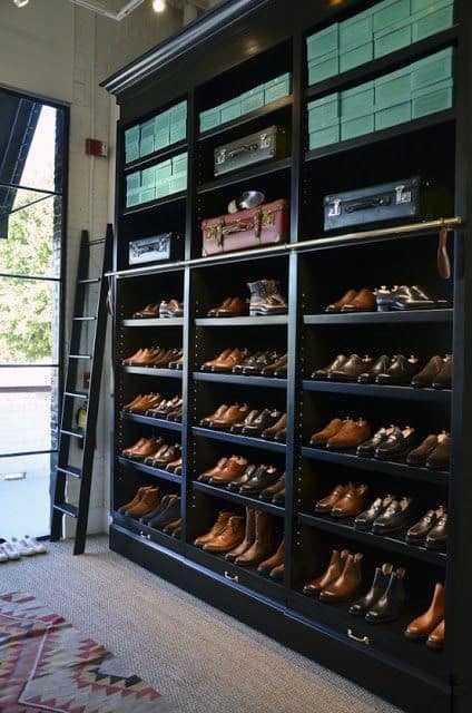 Shoe display with brown shoes, green boxes, and suitcases on a black shelf next to a ladder in a boutique