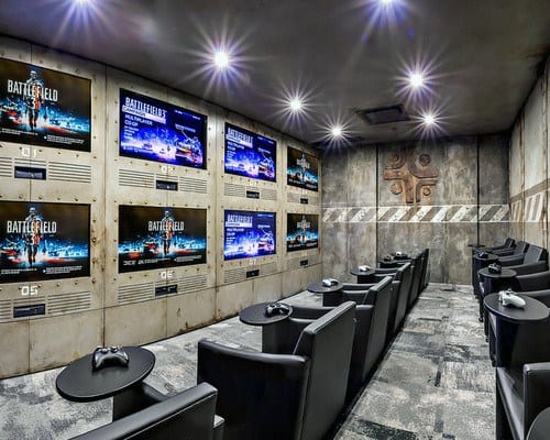 Gaming room with multiple screens showing video game graphics and rows of black chairs with controllers