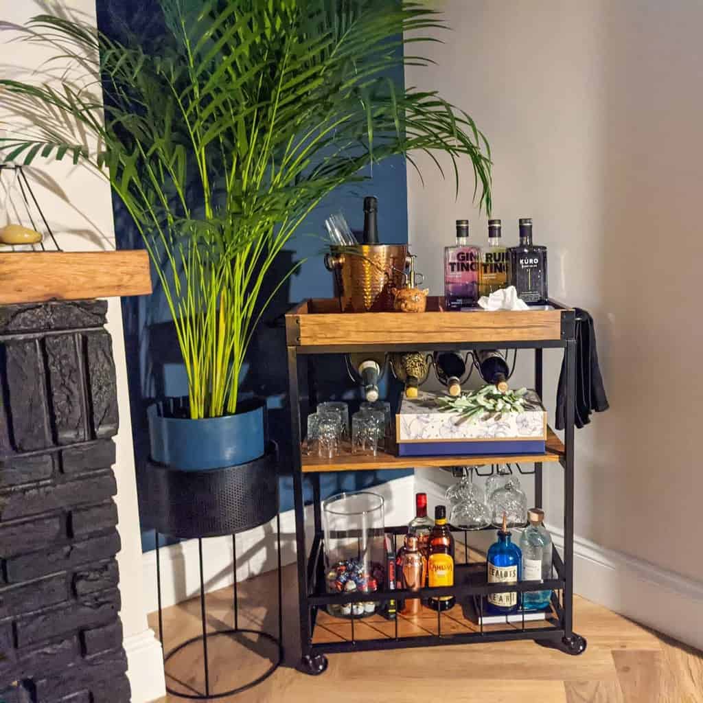 Wood and steel bar cart with liquor bottles, wine, glassware, and a potted plant nearby.