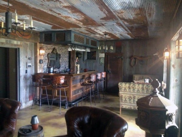 Rustic industrial-style mancave with distressed metal ceiling, wooden bar, leather chairs, and brick accents.