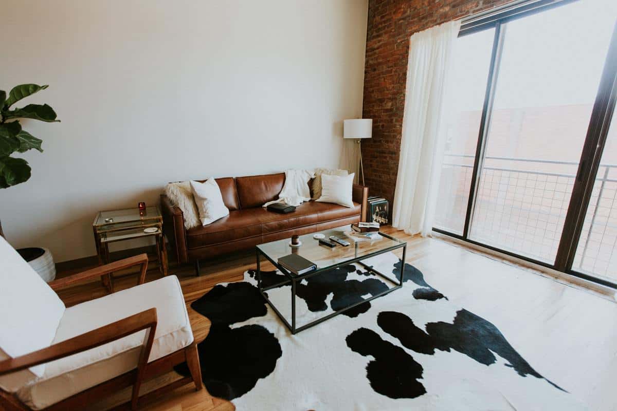 Modern living room with a rich brown leather sofa, glass coffee table, cowhide rug, and large window