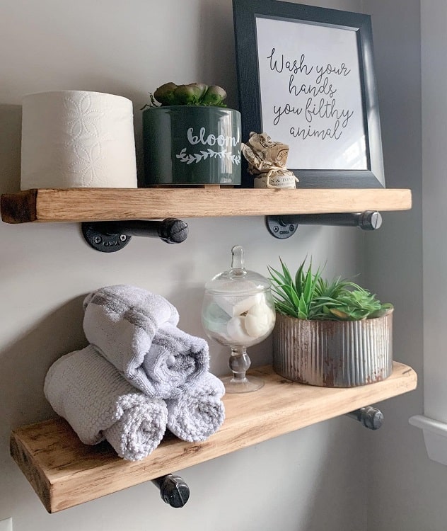 Industrial farmhouse bathroom shelves with rolled towels, potted plants, and framed wall decor.