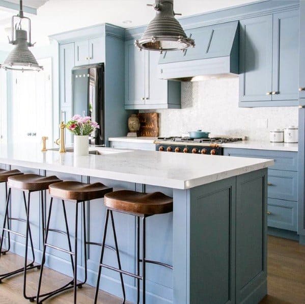 Charming kitchen with industrial-style large pendant lights above a white marble island, accented with blue cabinetry and wooden bar stools