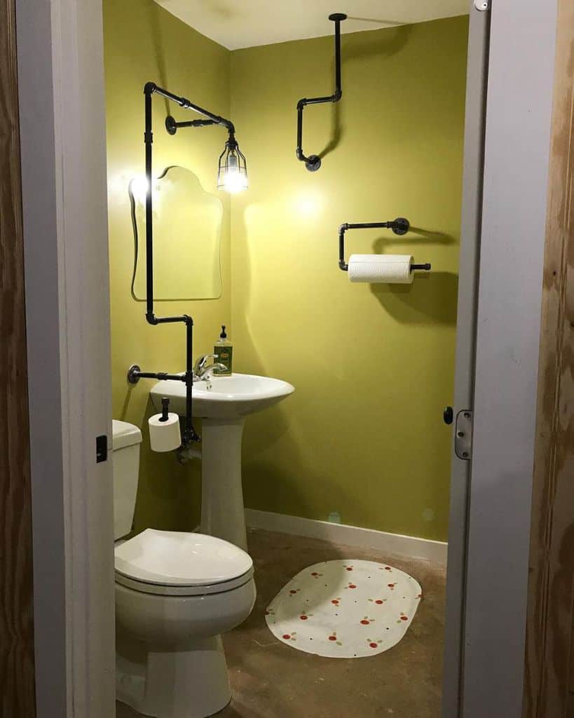 Industrial-style bathroom with exposed black pipes, pedestal sink, and green walls.