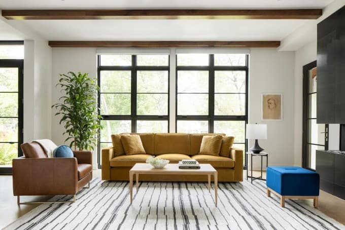 Modern living room with mustard sofa, leather armchair, striped rug, and large black-framed windows.