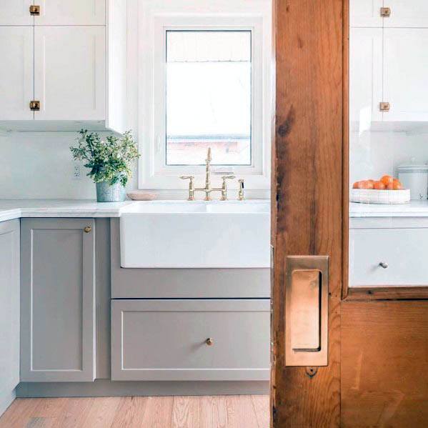 Warm wood pocket door with brass hardware opens to a bright farmhouse kitchen featuring white cabinetry, a farmhouse sink, and gold fixtures