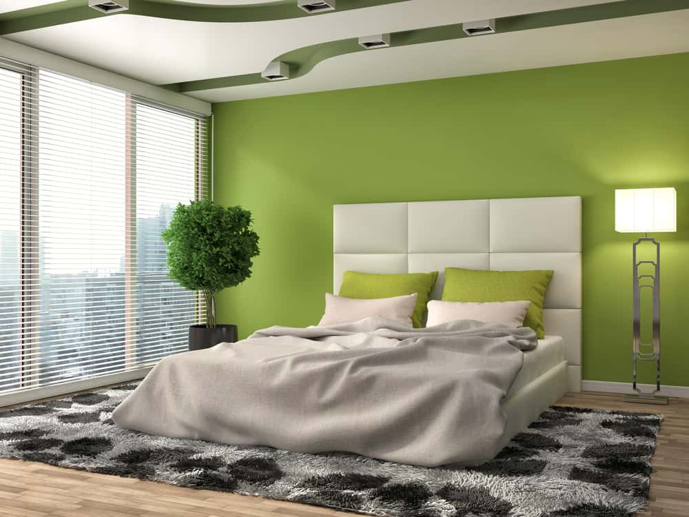 Modern bedroom with a green accent wall, white bed, green pillows, rug, tall plant, and floor lamp