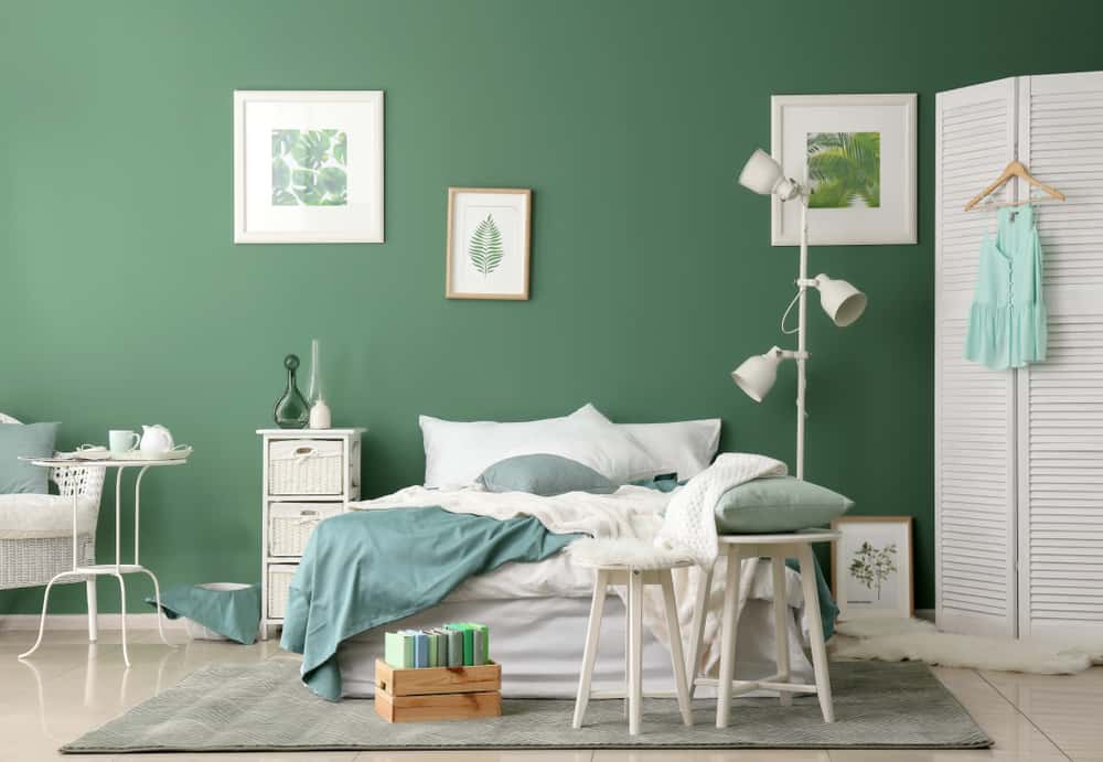 A green-themed bedroom with a bed, white lamp, and framed art on a green wall. A chair holds books and a cushion